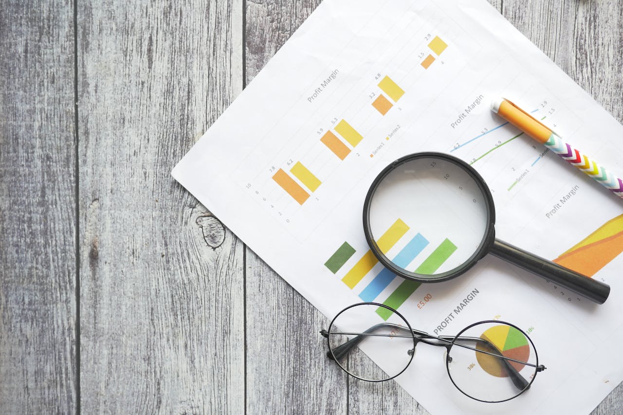 about-02 Overhead view of financial charts, a magnifying glass, ballpen, and eyeglasses on a wooden surface.