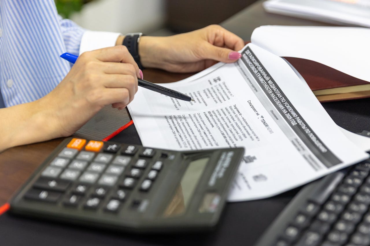 about-img Close-up of a person analyzing financial documents using a calculator and pen.